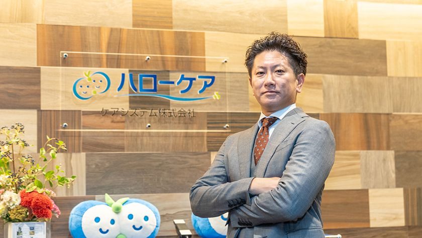 ハローケアのロゴの前に立つ代表取締役 加藤一朗